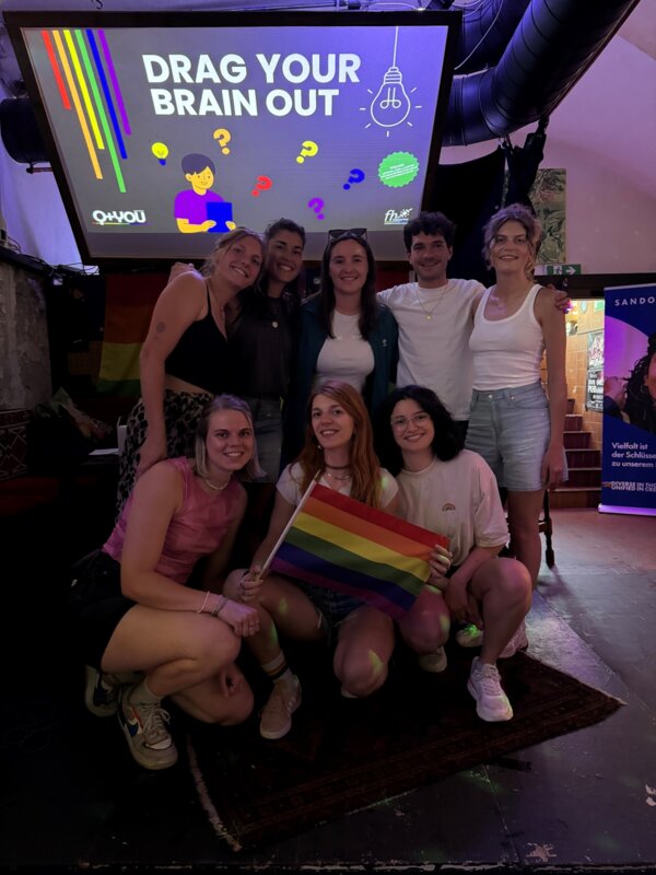 Eine Gruppe von acht Studierenden posiert lächelnd vor der Leinwand mit der Aufschrift „Drag Your Brain Out“ beim Pub Quiz des Praxisprojekts QYOU an der HOK. Eine Person hält eine Regenbogenflagge, im Hintergrund sind Regenbogenfarben und Symbole des Quizdesigns zu sehen.