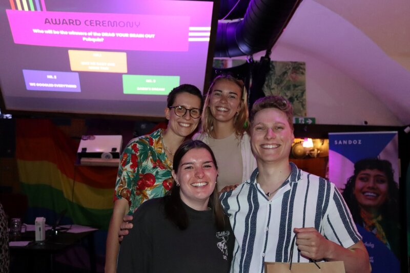 Vier Studierende des Gewinnerteams posieren lachend bei der Award Ceremony des Pub Quiz „Drag Your Brain Out“ im Rahmen des Praxisprojekts QYOU an der HOK. Im Hintergrund ist die Leinwand mit der Ankündigung der Siegerehrung zu sehen.