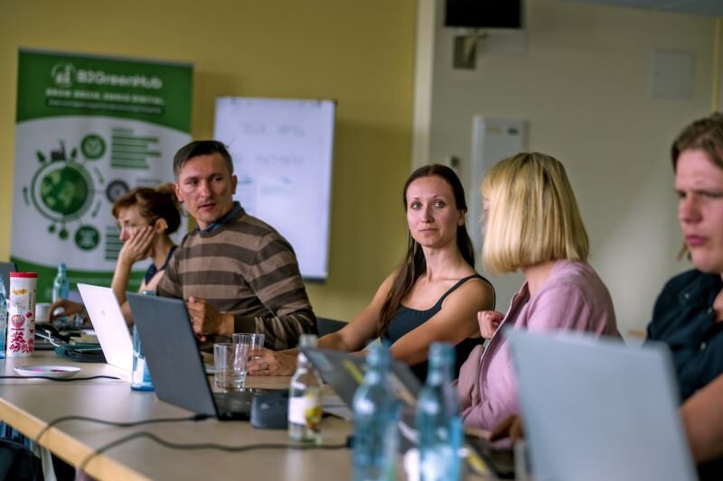 Mehrere Teilnehmende des GREENE4-Partnertreffens in Bautzen sitzen an einem Konferenztisch mit Laptops und Getränken, während sie konzentriert einer Diskussion folgen. Im Hintergrund ist ein Banner des Projekts zu sehen.