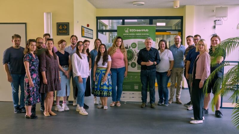 Gruppenfoto des GREENE4.0-Projektteams beim Partnertreffen in Bautzen. Rund 15 Personen stehen in einem Flur neben einem grünen Roll-Up mit dem Logo und den Inhalten des Projekts B2GreenHub.
