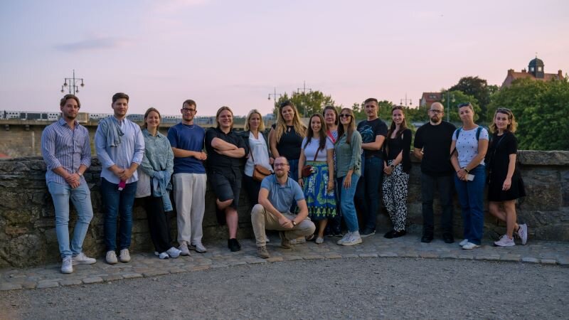 Gruppenfoto der GREENE4-Projektgruppe beim Partnertreffen in Bautzen. Die Teilnehmenden stehen im Freien an einer Steinmauer mit Blick auf Bäume und Gebäude im Hintergrund.