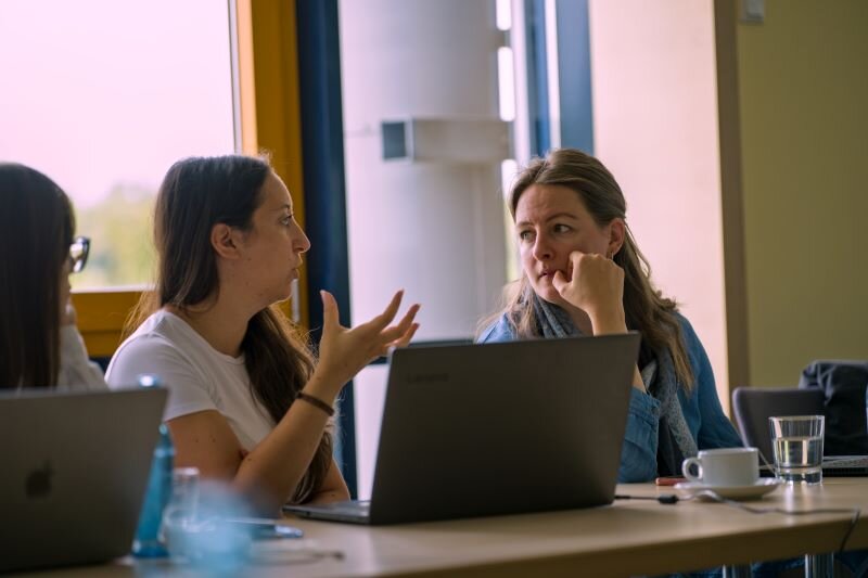 Zwei Teilnehmerinnen des GREENE4-Projekts im Gespräch während des Partnertreffens in Bautzen. Beide sitzen an einem Tisch mit Laptops, Wasser und Kaffeetasse und diskutieren angeregt.