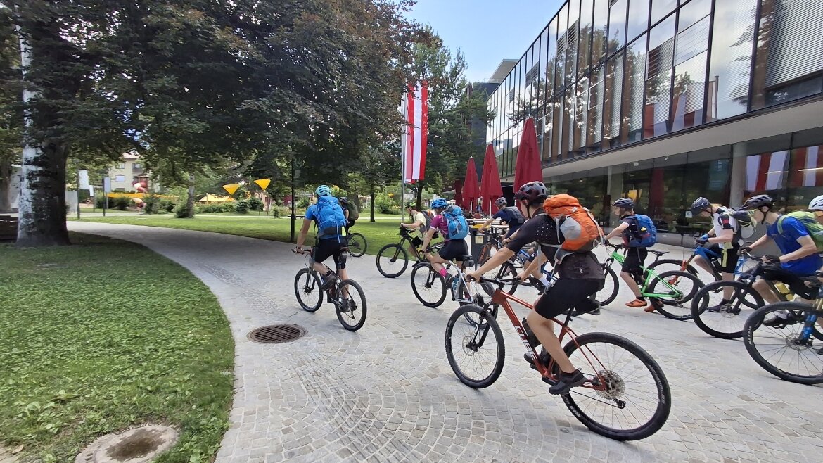 Mehrere Schülerinnen und Schüler auf Mountainbikes fahren vom Campus der HOK los. Im Hintergrund sind das moderne Hochschulgebäude, rote Sonnenschirme und österreichische Fahnen zu sehen.