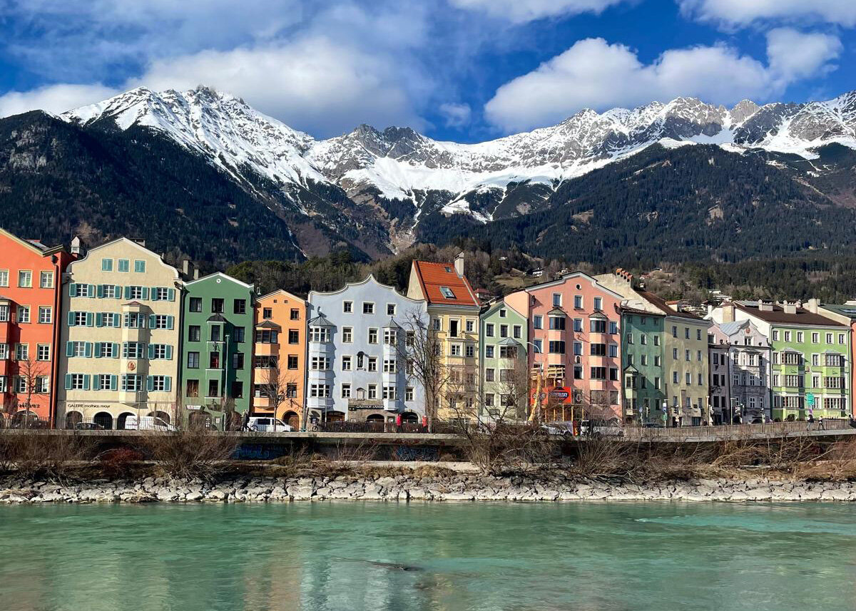Bunte Häuserreihe am Ufer des Inns in Innsbruck mit schneebedeckten Bergen der Nordkette im Hintergrund unter blauem Himmel.
