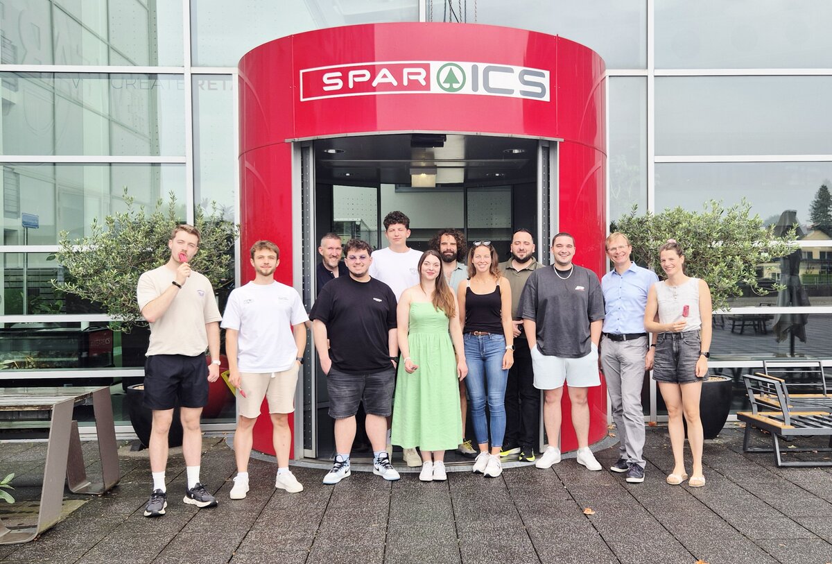 Eine Gruppe von Studierenden und Lehrenden steht lächelnd vor dem auffälligen roten Eingangsbereich der SPAR ICS in Salzburg. Einige genießen ein Eis, was der Szene eine sommerlich-lockere Stimmung verleiht.