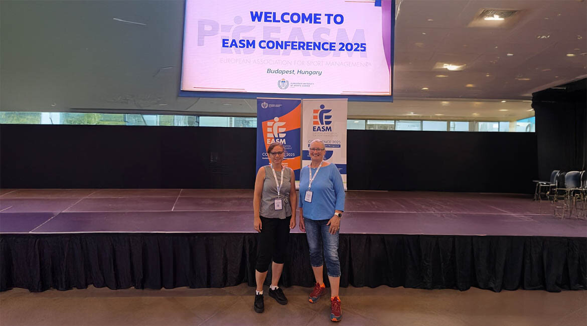 Zwei Teilnehmerinnen der HOK stehen lächelnd vor einer Bühne mit EASM-Banner und Konferenzschild bei der EASM Conference 2025 in Budapest, Ungarn.