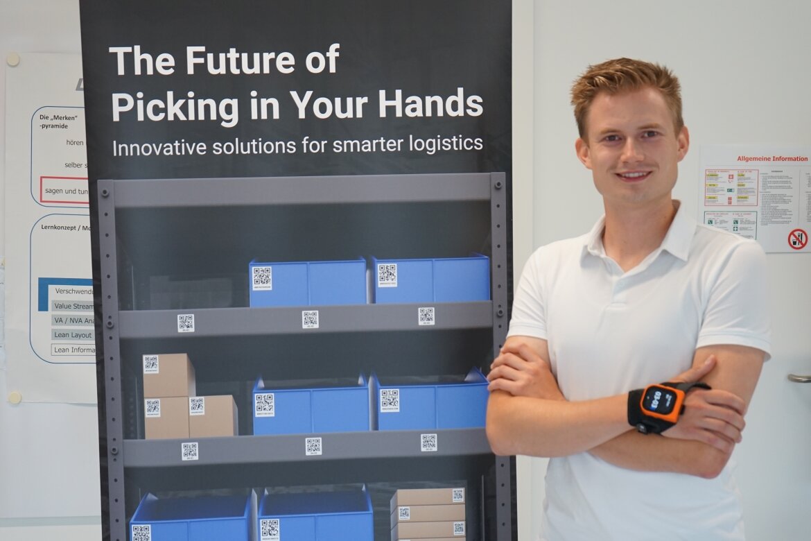 Ein junger Mann in weißem Poloshirt steht mit verschränkten Armen vor einem Roll-up mit dem Titel „The Future of Picking in Your Hands“. Am Handgelenk trägt er einen ProGlove-Scanner.