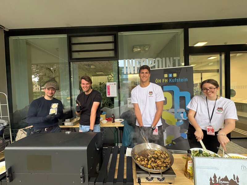 Vier junge Menschen stehen an einem Kochstand vor dem Infopoint der HOK und bereiten gemeinsam Gemüsegerichte zu. Im Hintergrund ist ein ÖH-Banner sichtbar.