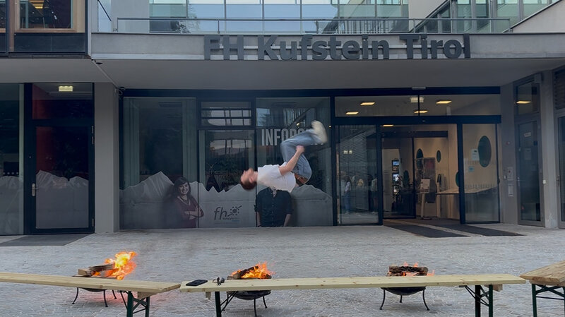 Ein junger Mann macht einen Rückwärtssalto zwischen zwei Feuerschalen vor dem Haupteingang der HOK. Im Hintergrund ist das Gebäude mit Glasfassade und das FH-Logo zu sehen.