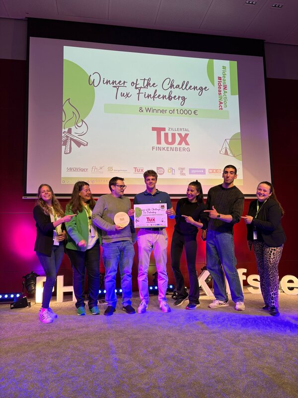 Sieben Personen posieren auf einer Bühne mit einem Siegerscheck und einer Holztrophäe. Im Hintergrund ist auf der Leinwand der Schriftzug „Winner of the Challenge Tux Finkenberg & Winner of 1.000 €“ zu sehen.