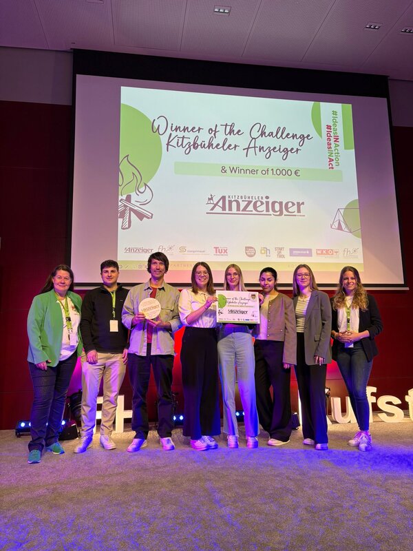 Eine Gruppe von neun Personen steht auf der Bühne und hält ein Zertifikat sowie einen Preis. Auf der Leinwand im Hintergrund steht: „Winner of the Challenge Kitzbüheler Anzeiger & Winner of 1.000 €“.
