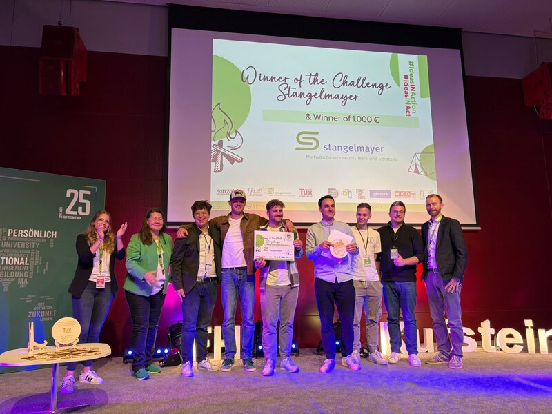 Elf Personen stehen auf einer Bühne und feiern gemeinsam. In der Mitte hält ein Team ein Zertifikat und eine Trophäe. Auf der Leinwand steht: „Winner of the Challenge Stangelmayer & Winner of 1.000 €“.