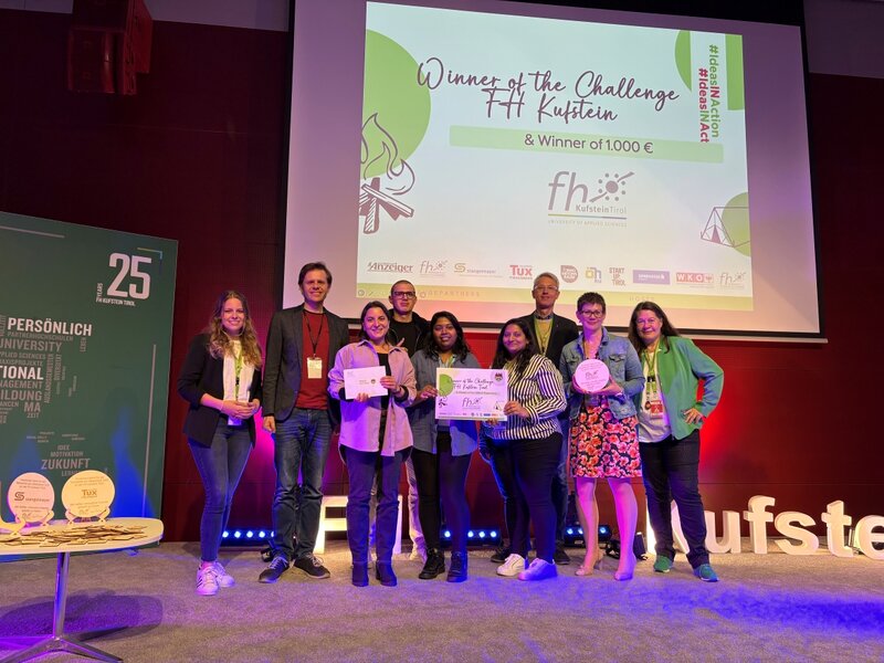 Eine Gruppe von neun Personen steht auf der Bühne und hält Urkunden sowie einen Preis in Händen. Im Hintergrund ist eine Leinwand mit der Aufschrift „Winner of the Challenge HOK & Winner of 1.000 €“ zu sehen.