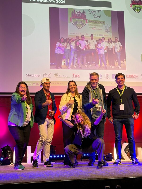 Eine Gruppe von sechs Personen posiert energiegeladen auf der Bühne des INNoCamp 2025 vor einem großen Bildschirm mit Gruppenfoto und Logos der Partnerorganisationen.