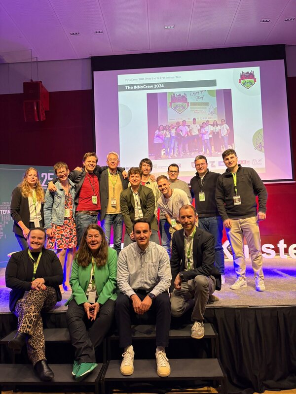 Gruppenfoto von rund 16 Personen auf einer Bühne vor einer Leinwand. Die Teilnehmer:innen der INNoCrew 2024 posieren lachend, einige stehen, andere sitzen auf den Stufen. Im Hintergrund ist ein Bild mit dem Text „The INNoCrew 2024“ projiziert.