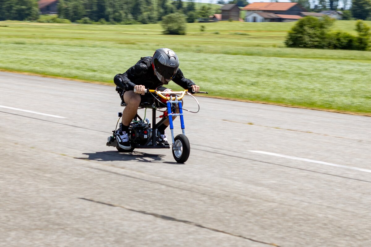 Ein junger Mann mit Schutzkleidung und Helm fährt in geduckter Haltung ein selbstgebautes Mini-Bike auf einer Landstraße. Das Fahrzeug ist extrem klein und liegt tief über dem Asphalt.