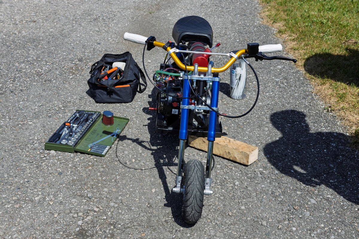 Frontale Nahaufnahme eines kleinen Mini-Bikes mit sichtbarem Motor, goldfarbenem Lenker und blauen Gabelrohren. Daneben liegen Werkzeugkoffer, ein Luftfilter und eine Tasche mit Schraubwerkzeug auf dem Asphalt.