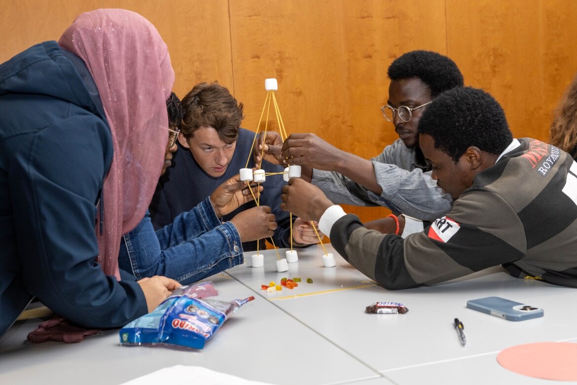 Eine diverse Gruppe Studierender baut gemeinsam eine Struktur aus Spaghetti und Marshmallows im Rahmen eines Workshops zu Teamarbeit und Problemlösung.