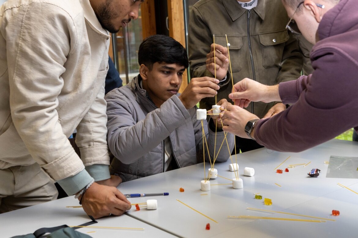 Eine Gruppe von Studierenden konstruiert konzentriert einen Turm aus Spaghetti und Marshmallows im Rahmen eines interaktiven Workshops.