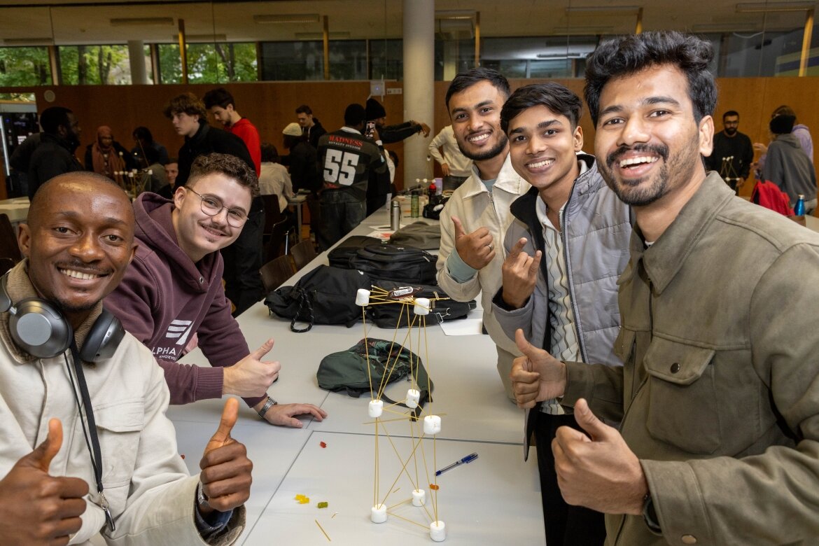Eine Gruppe lachender Studierender zeigt stolz ihren fertigen Turm aus Marshmallows und Spaghetti im Workshop.
