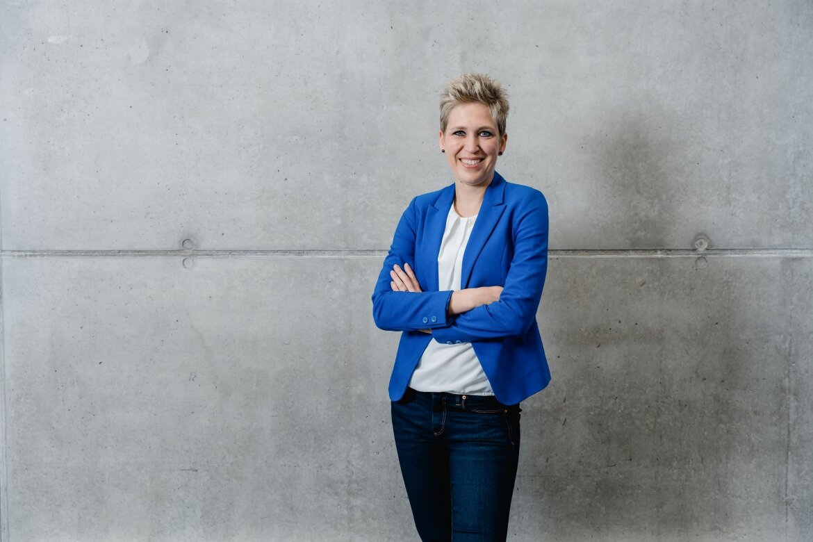 Kristina Kampfner steht lächelnd mit verschränkten Armen vor einer grauen Betonwand, gekleidet in einem blauen Blazer und weißer Bluse.