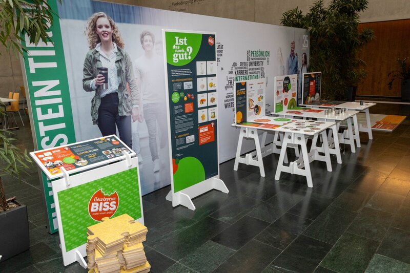 Verschiedene interaktive Stationen der Ausstellung „Gewissensbiss“ in der Aula der HOK. Die Tafeln und Tische informieren spielerisch über Lebensmittelverschwendung, Haltbarkeit und Geschmack.