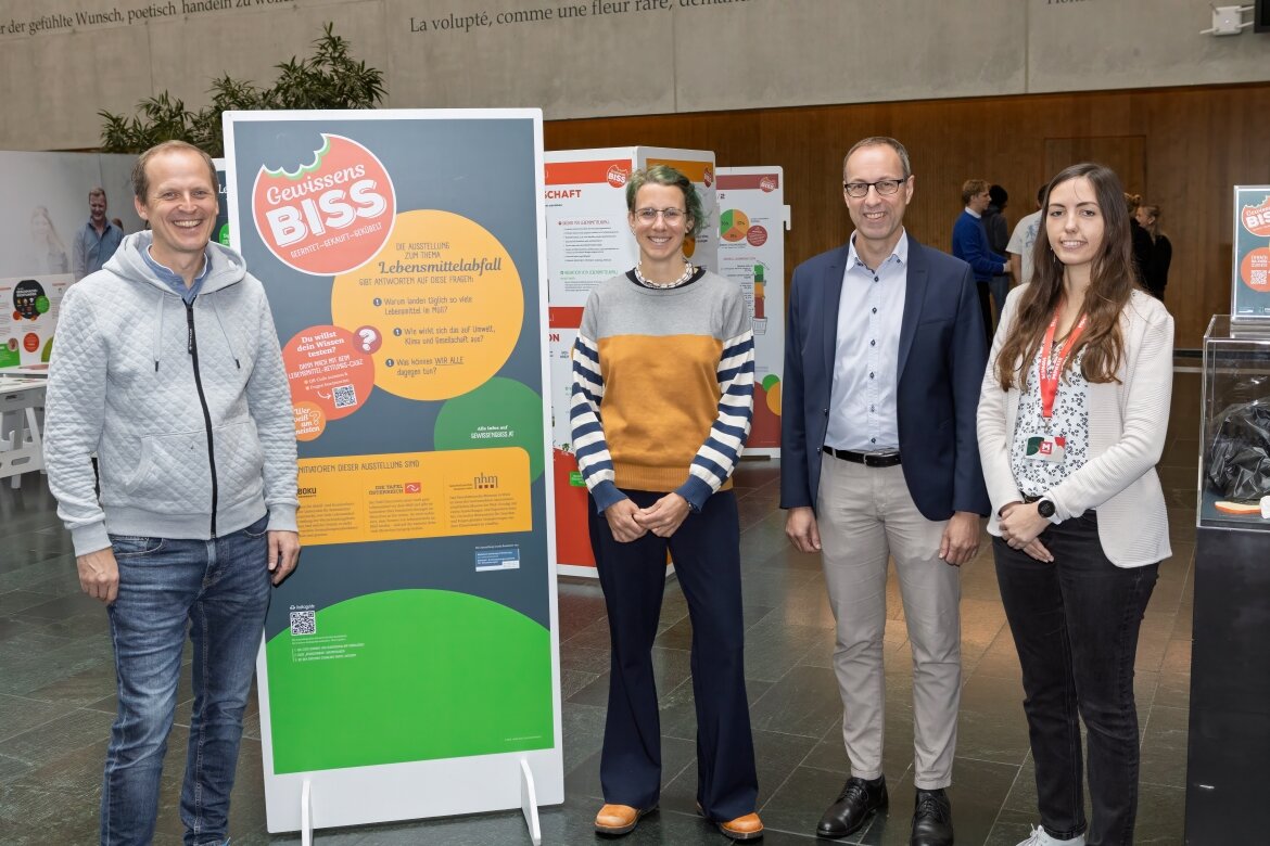 Vier Personen stehen neben einem großen Informationsplakat zur Ausstellung „Gewissensbiss“ in der Aula der HOK. Die Ausstellung thematisiert Lebensmittelabfall und nachhaltigen Konsum. Im Hintergrund sind weitere interaktive Stationen sichtbar.