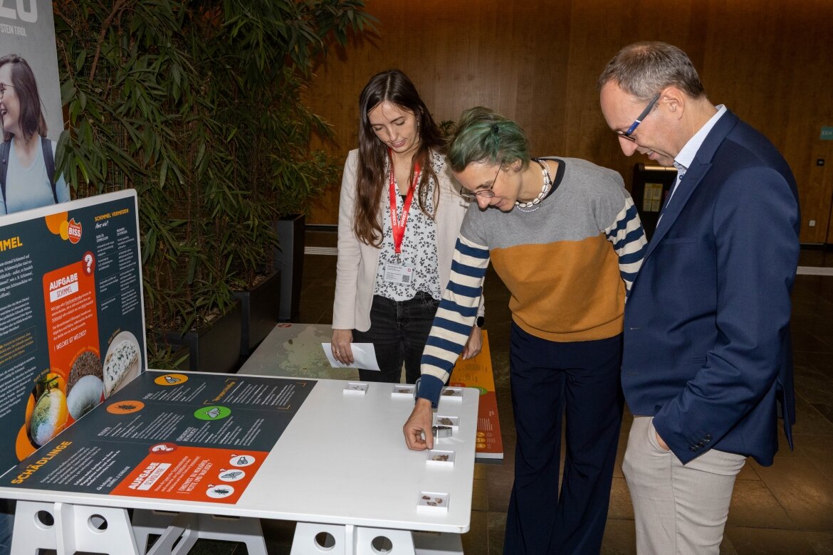 Drei Personen interagieren mit einer Mitmachstation der Ausstellung „Gewissensbiss“ in der Aula der HOK. Die Station beschäftigt sich mit dem Thema Schimmel und Schädlingsbefall bei Lebensmitteln.