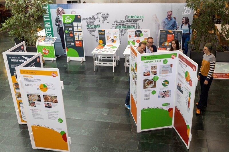 Blick von oben auf die Ausstellung „Gewissensbiss“ in der Aula der HOK. Die Ausstellung umfasst Informationstafeln und Mitmachstationen zu Themen wie Lebensmittelabfall, Konsum und Haltbarkeit. Einige Besucher:innen betrachten interaktiv die Inhalte.
