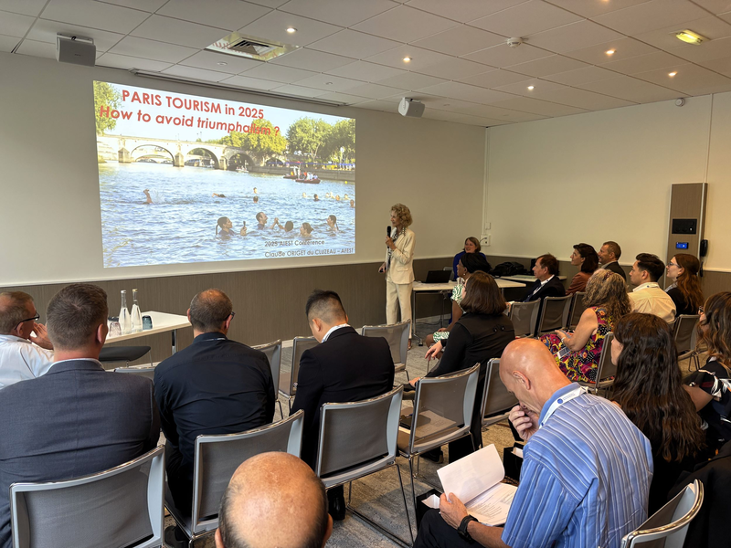 Claude Origet du Cluzeau hält einen Vortrag bei der AIEST-Jahrestagung 2025 zum Thema „Paris Tourism in 2025 – How to avoid triumphalism?“. Im Konferenzraum folgen zahlreiche Teilnehmende aufmerksam der Präsentation, die ein Bild von Badenden in der Seine zeigt.