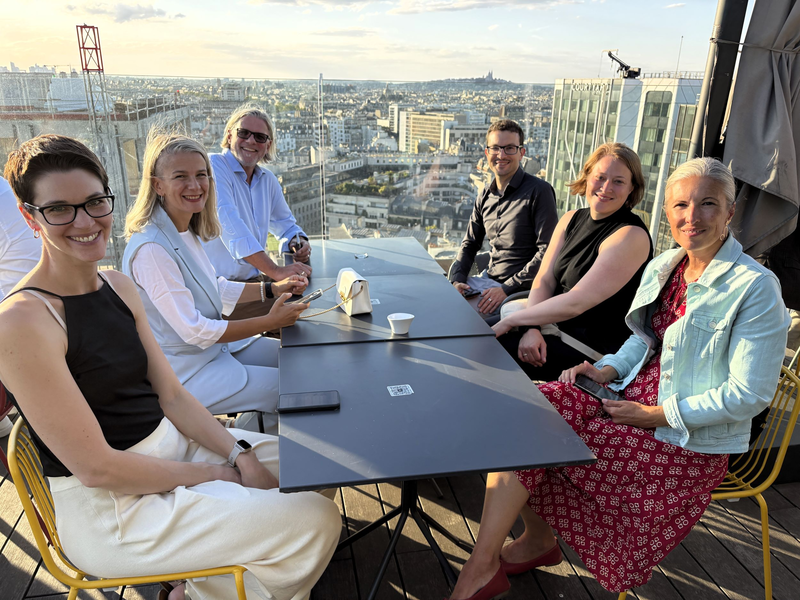 Sechs Personen sitzen in entspannter Atmosphäre auf einer Rooftop-Bar bei Sonnenuntergang. Im Hintergrund ist die Skyline von Paris mit Blick auf die Basilika Sacré-Cœur zu sehen.