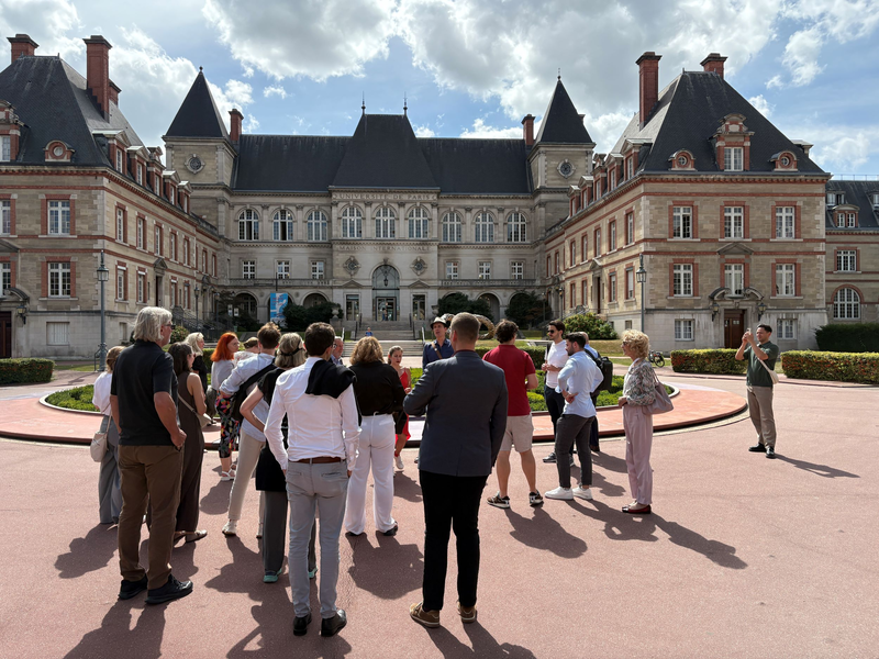 Eine Gruppe von Konferenzteilnehmenden steht vor dem imposanten Gebäude der Maison Internationale in Paris. Die historische Fassade mit Türmen und roten Backsteinelementen ist bei sonnigem Wetter gut sichtbar.