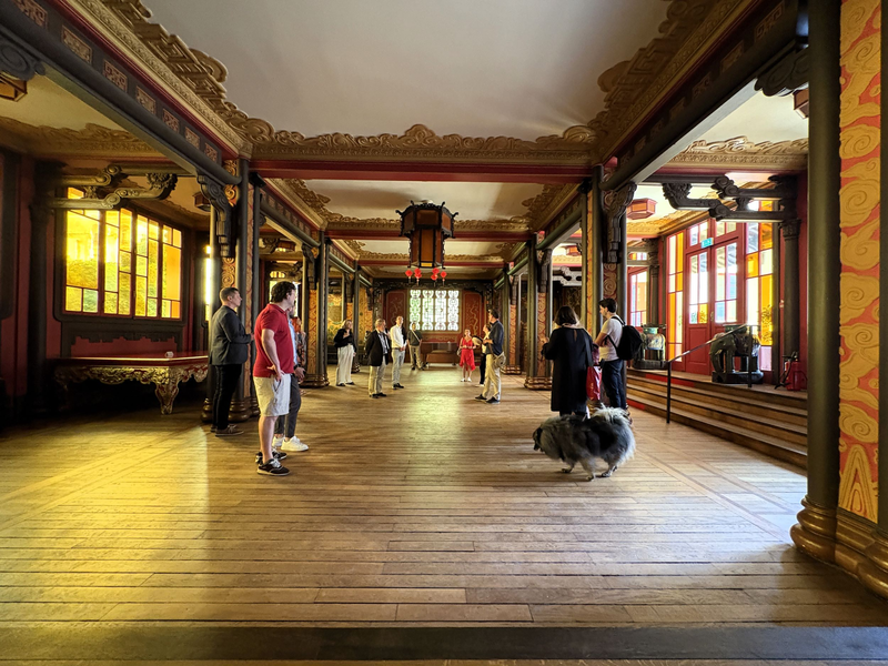 Mehrere Personen stehen in einem reich verzierten Saal mit asiatischer Architektur im Cité Internationale Universitaire de Paris. Die farbigen Fenster und Holzverzierungen schaffen eine besondere Atmosphäre. Ein Hund läuft durch das Bild.