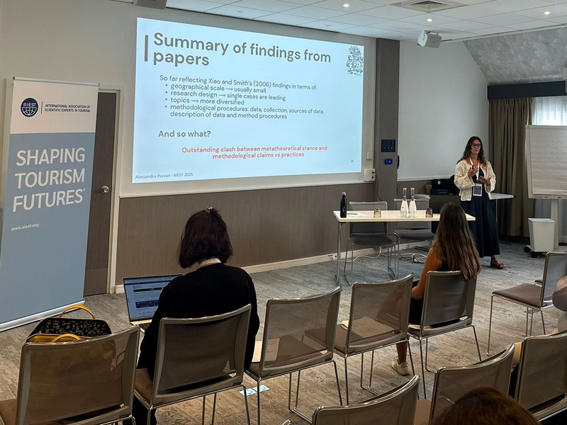 Alessandra Pozzan steht neben einer Präsentationsleinwand und hält einen Vortrag. Die Folie zeigt eine Zusammenfassung wissenschaftlicher Erkenntnisse. Zwei Zuhörerinnen sitzen im Raum. Links im Bild steht ein Roll-Up mit dem AIEST-Logo und dem Slogan „Shaping Tourism Futures“.