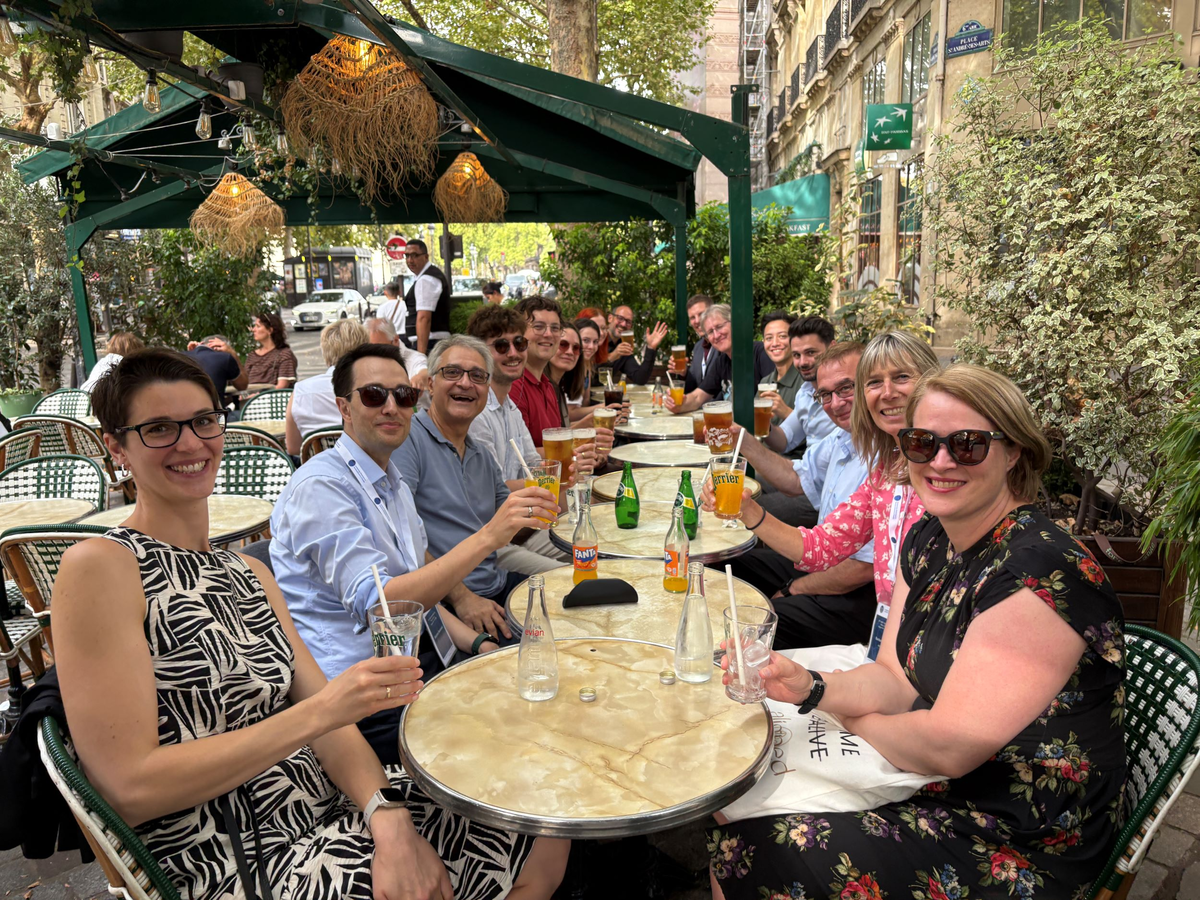 Eine große Gruppe von Konferenzteilnehmenden sitzt fröhlich an einem langen Tisch in einem Straßencafé, hebt die Gläser und genießt einen gemeinsamen Apéritif zum Abschluss der AIEST-Jahrestagung 2025.