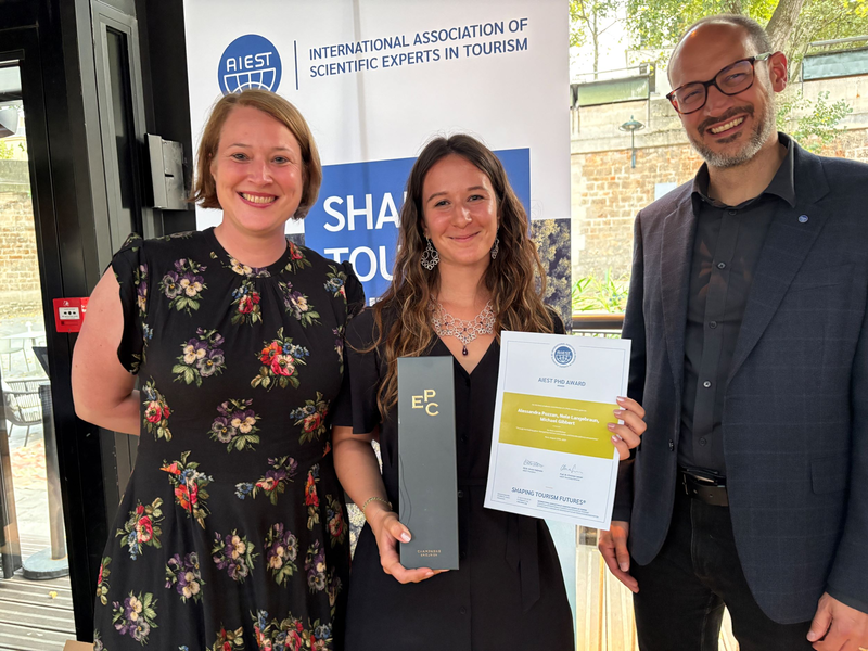 Drei Personen stehen lächelnd nebeneinander, während Alessandra Pozzan in der Mitte stolz den AIEST PhD Award 2025 sowie eine Urkunde in den Händen hält. Im Hintergrund ist ein Banner der AIEST zu sehen.