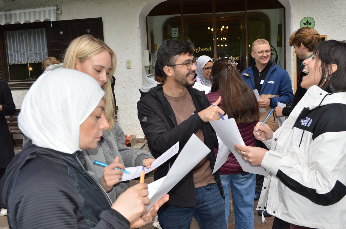 Gruppe von Studierenden steht lachend zusammen vor dem Berghotel Hinterduxerhof und füllt Arbeitsblätter aus.