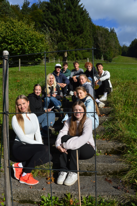 Gruppe von Studierenden sitzt auf einer Wiese am Hang in der Sonne und lächelt in die Kamera.