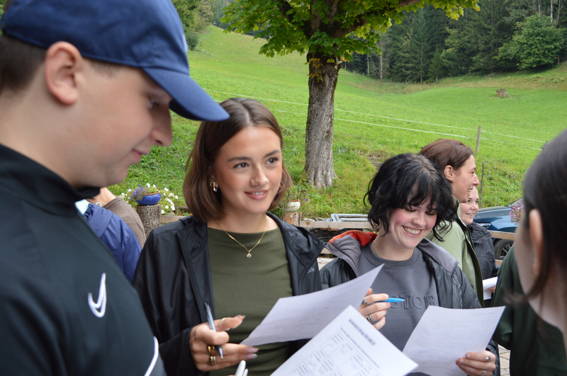 Mehrere Studierende stehen im Freien, halten Blätter in den Händen und lachen während einer Kennenlernaktivität am Hinterduxerhof.