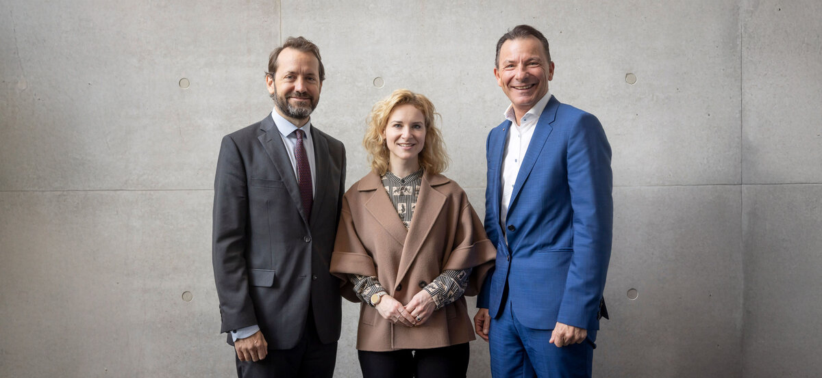 Drei Personen stehen nebeneinander vor einer grauen Wand.
