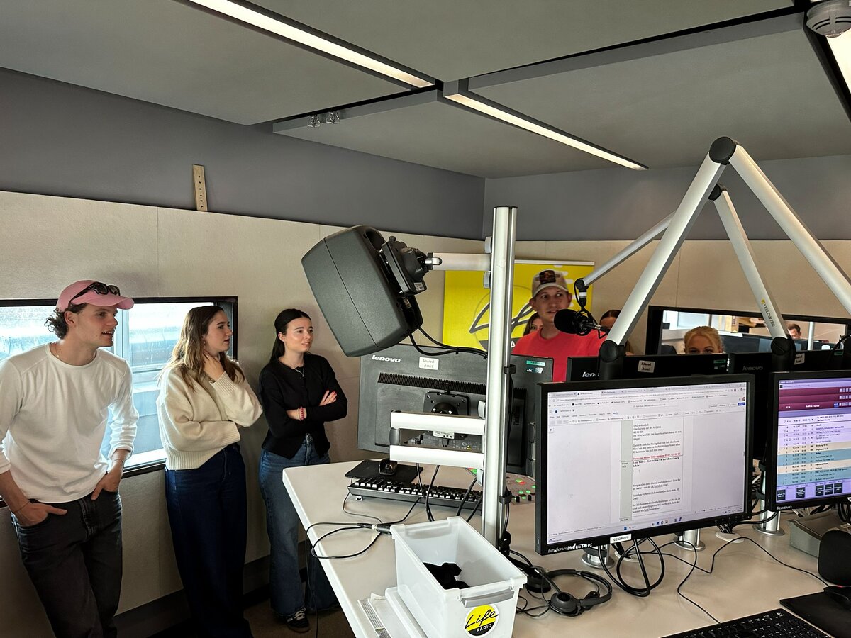 Studierende stehen im Radiostudio zwischen Mikrofonen, Monitoren und Studiotechnik und hören den Erklärungen eines Radioprofis zu.