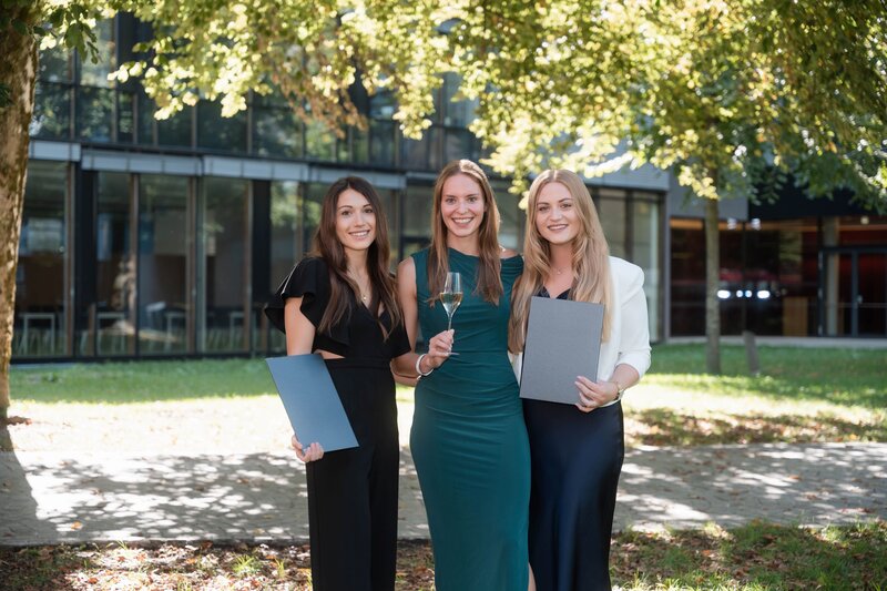 Drei Absolvierende posieren für ein Gruppenfoto auf dem Campus vor dem HOK-Gebäude.