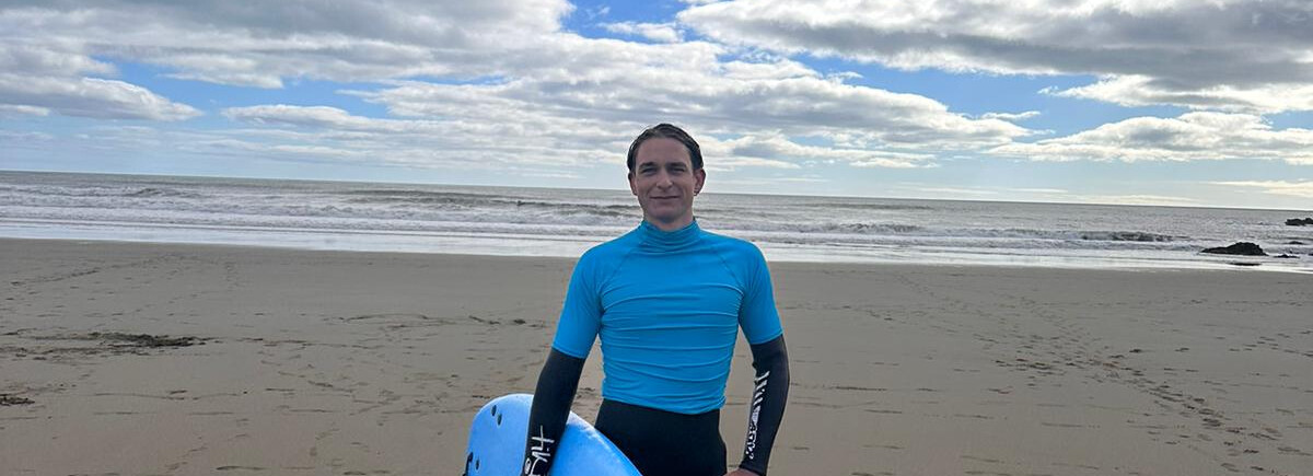 Ein junger Mann in einem Neoprenanzug und blauem Shirt mit einem Surfboard am Strand.