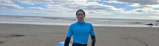 Ein junger Mann in einem Neoprenanzug und blauem Shirt mit einem Surfboard am Strand.